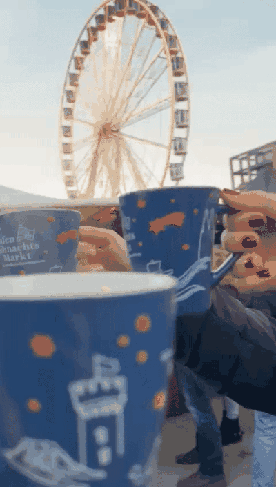 Hands holding blue Christmas-market mugs with a Ferris wheel behind